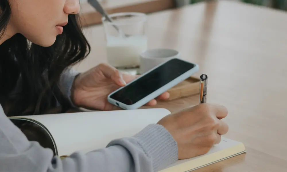 スマートフォンを見ながら英語学習ノートに書き込む女性。TOEIC対策を始めたばかりの様子