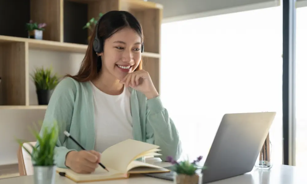 英語のシャドーイング練習をする女性。パソコンの前でヘッドホンをつけ、ノートと鉛筆を用意している。
